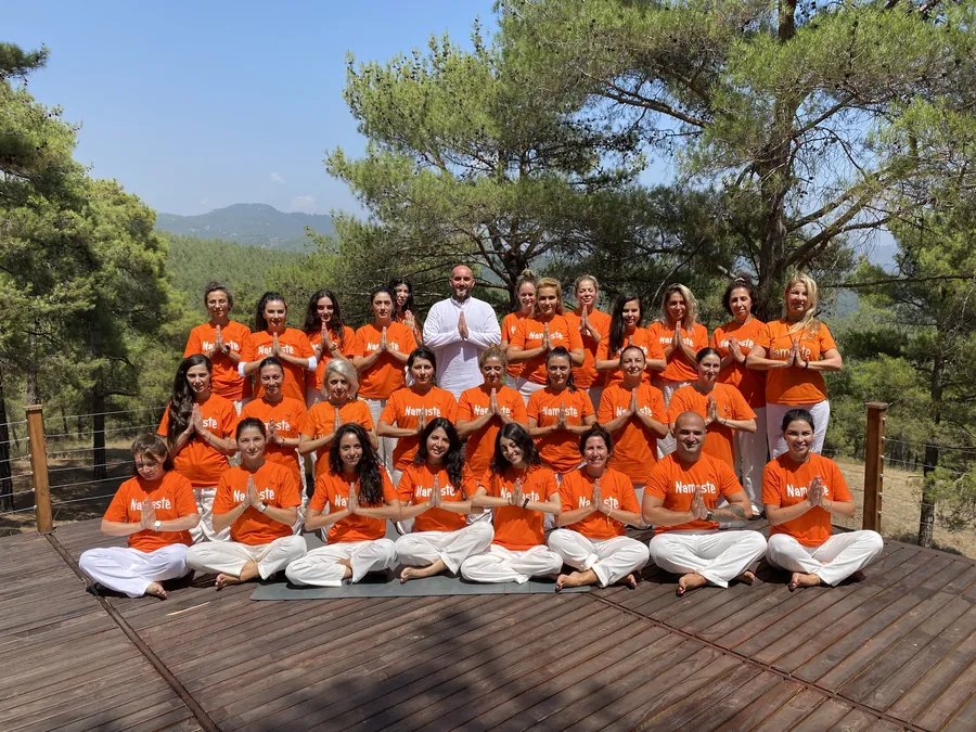 Ahşap terasta ellerini göğüste birleştirmiş toplu fotoğraf veren turuncu tişörtlü grup