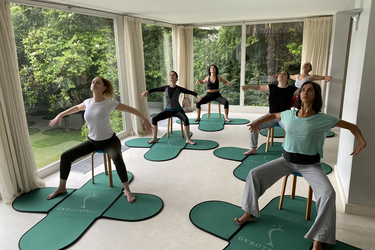 Cam duvarlı salonda sandalye ve Gyrotonic matlarıyla Gyrokinesis çalışması yapan grup